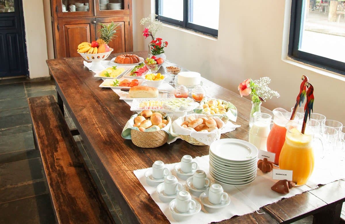 Bevor die Tierwelt ruft, sorgt das Lodge-Frühstück dafür, dass niemand hungrig aufbricht. Ein üppiges brasilianisches Frühstücksbuffet auf einem rustikalen Holztisch mit tropischen Früchten, Brötchen, Kuchen, Acerola-Saft, Maracuja-Saft und Espressotassen