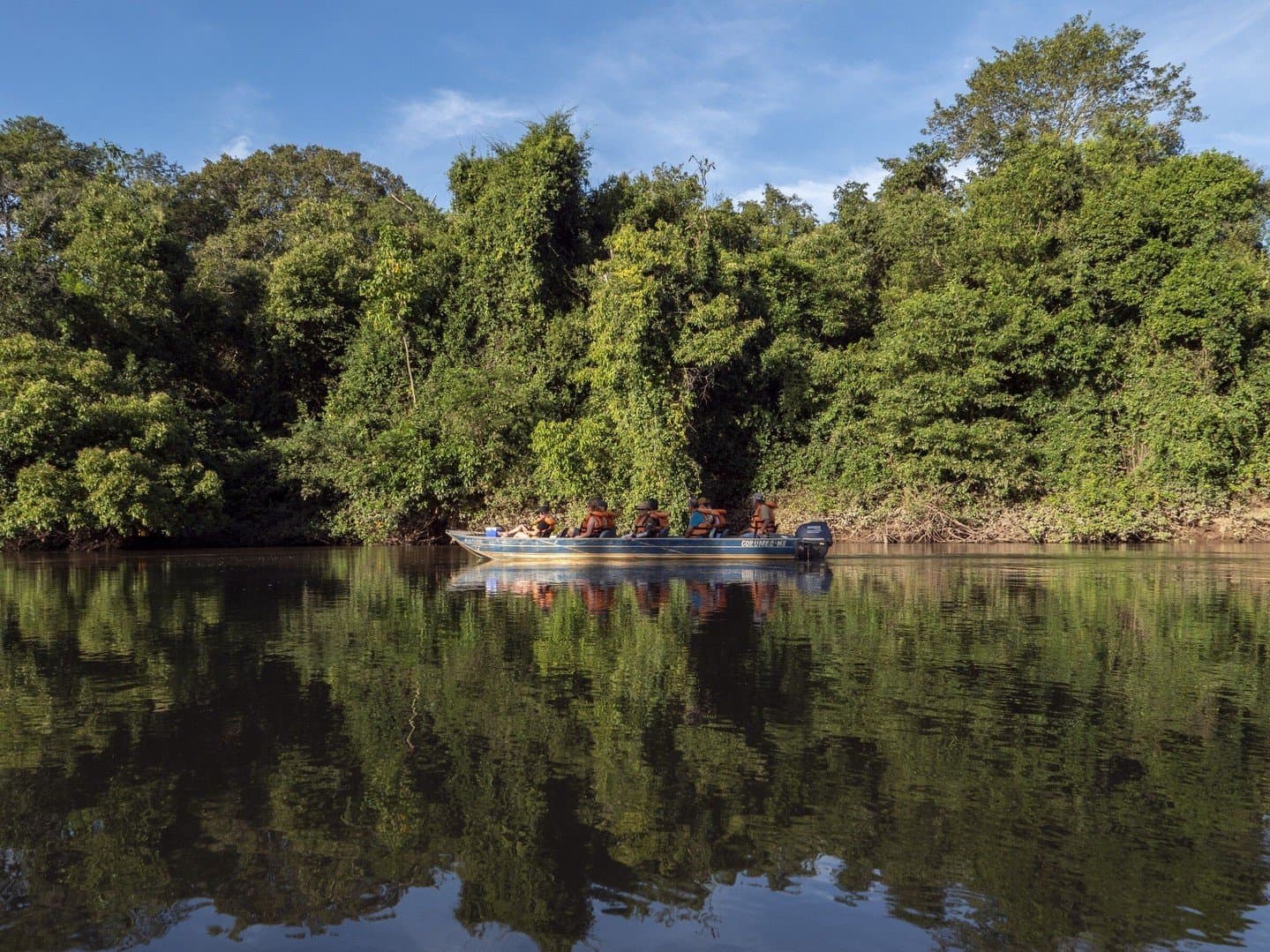 Stille auf dem Wasser, und der Wald rückt ganz nah heran. Ein kleines Motorboot mit Naturbeobachtern in orangefarbenen Schwimmwesten gleitet auf einem ruhigen Flusskanal entlang eines dichten grünen Auwaldes auf einer Pantanal-Bonito-Tour