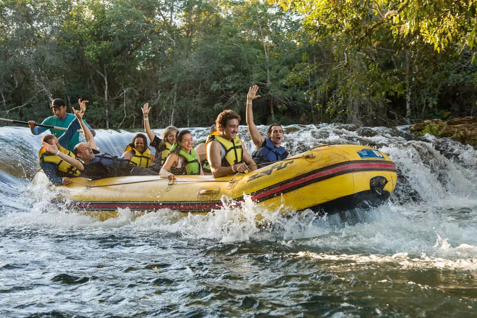Arme hoch, Kopf frei, Wasser überall, so sieht ein guter Nachmittag aus. Eine jubelnde Gruppe mit Schwimmwesten fährt auf einem gelben Schlauchboot durch schäumende Stromschnellen, umgeben von dichtem Galeriewald