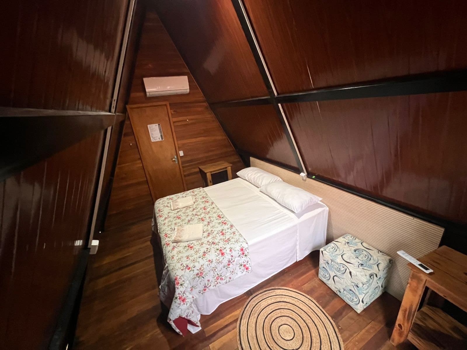 Simples, arrumado e aconchegante, do jeito que o Pantanal pede. Interior de chalé de madeira em formato A-frame com cama de casal, colcha florida, piso de madeira e ar-condicionado em um safari de onças no Pantanal econômico