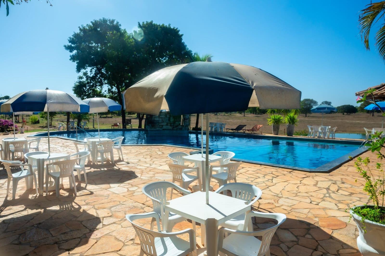 Entre uma saída de campo e outra, a piscina resolve bem. Área de piscina externa ensolarada com mesas brancas, cadeiras e guarda-sóis listrados em uma pousada de safari de onça no Pantanal