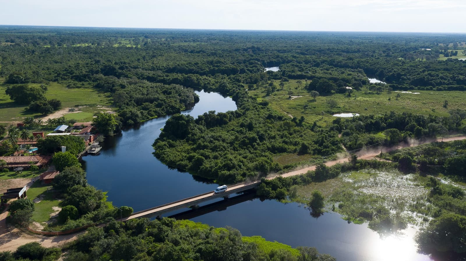 A ponte é pequena, mas o que fica do outro lado não tem tamanho. Vista aérea de uma van branca cruzando uma ponte de concreto sobre um rio sinuoso cercado pela densa vegetação úmida do Pantanal e uma pousada rústica à beira d'água