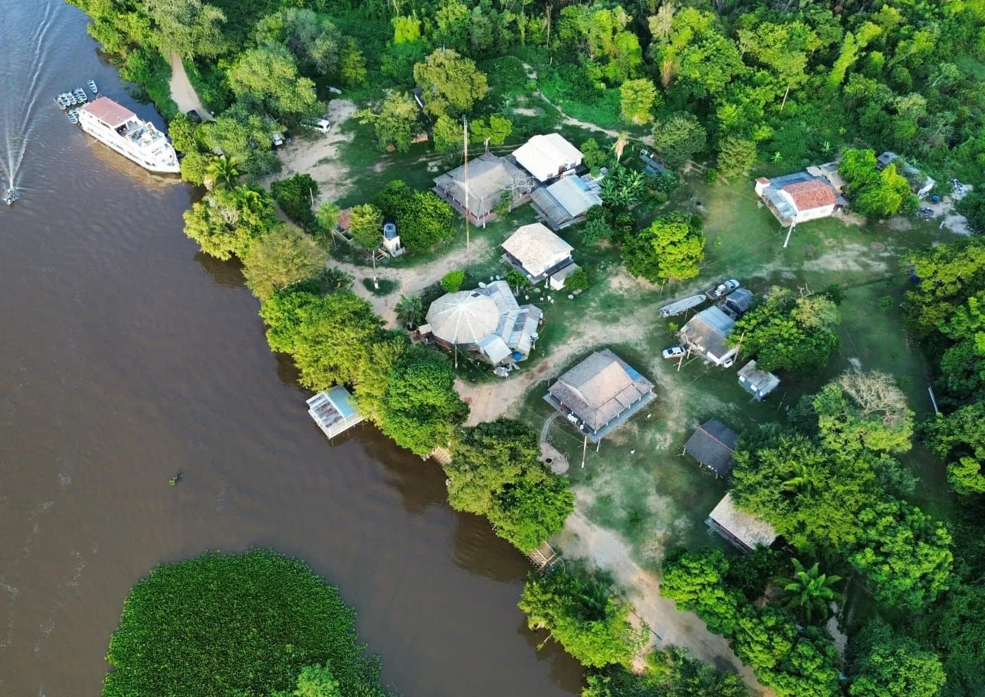 A pousada encostada no rio, do jeito que o Pantanal gosta. Vista aérea de drone de uma pequena pousada à beira do rio embutida em densa floresta tropical no Pantanal, com um barco de vários andares atracado na margem e construções com telhados de palha entre as árvores