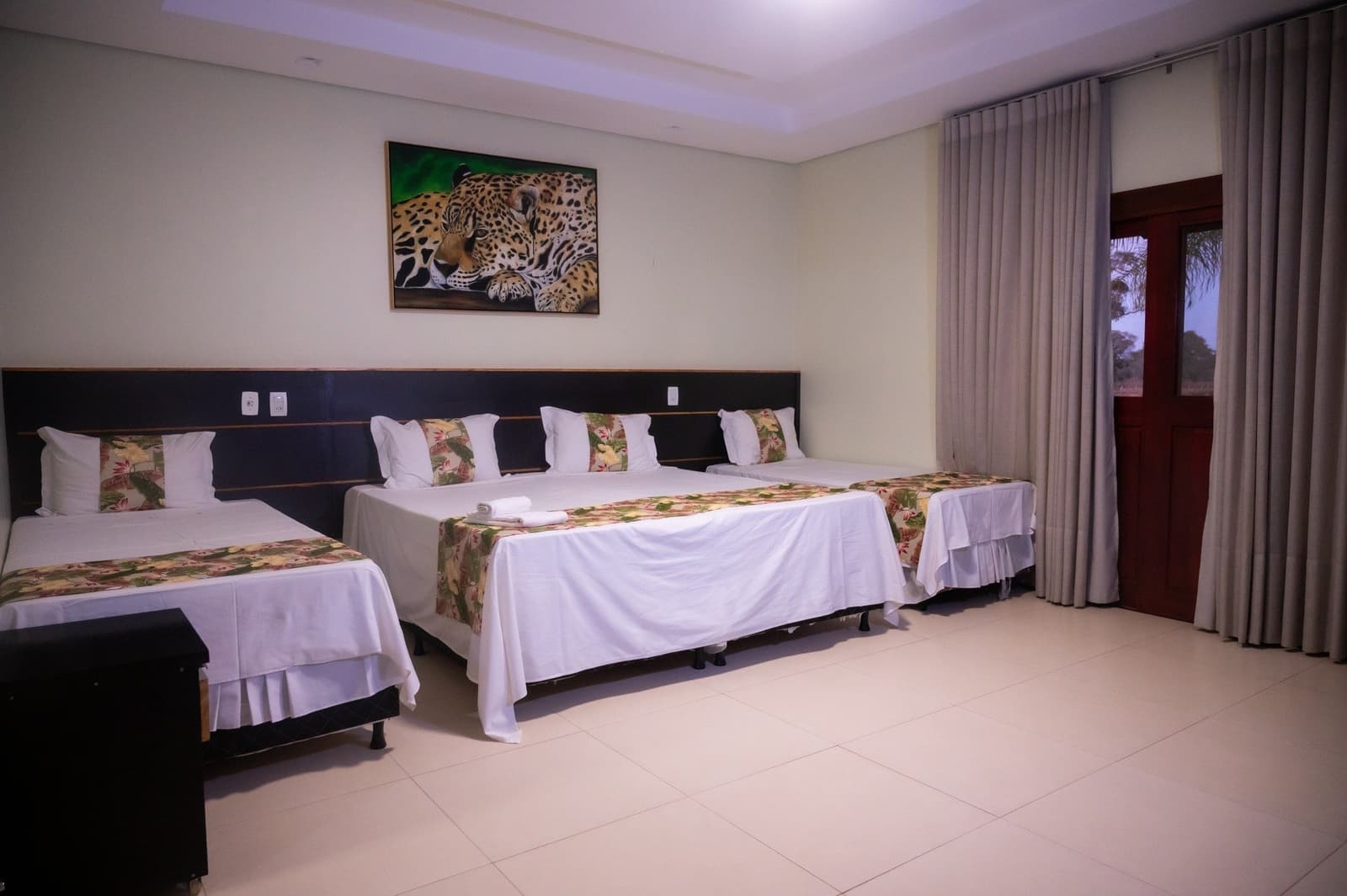 A onça da parede já te recebe antes de encontrar a de verdade. Quarto triplo espaçoso com roupas de cama brancas, pés de cama com estampa tropical e um quadro de onça-pintada acima da cabeceira escura