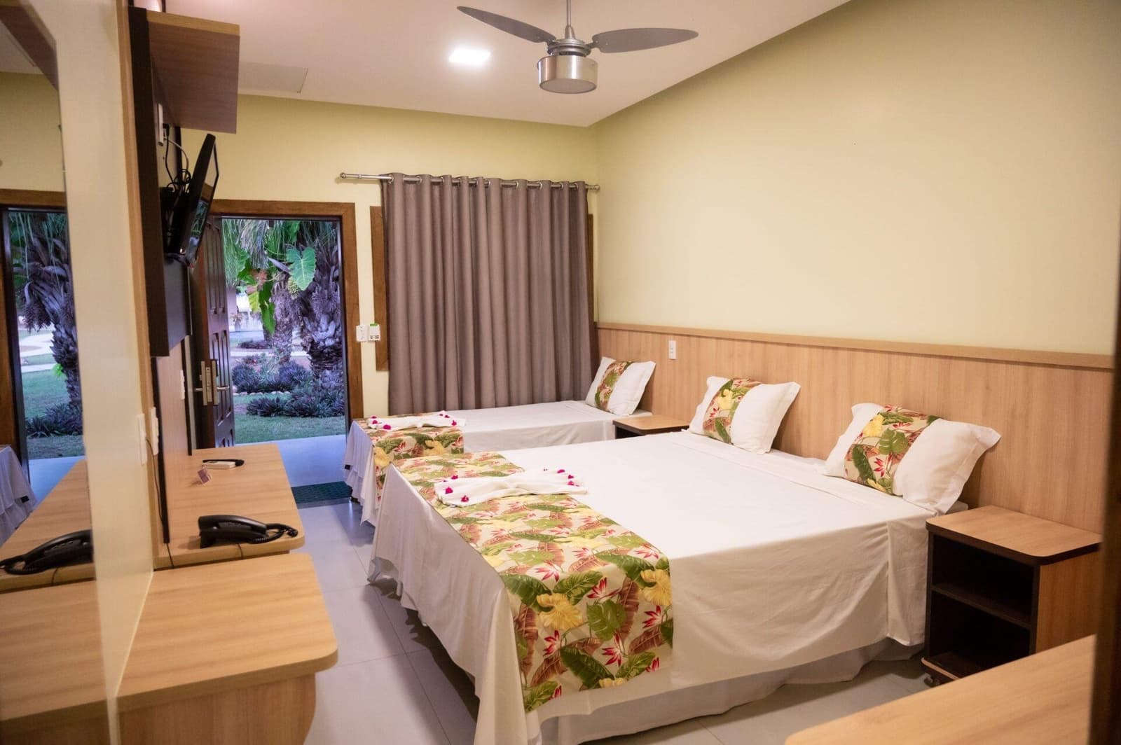 Confortável e aconchegante, do jeito certo para descansar entre as expedições. Quarto triplo em pousada de safari de jaguar no Pantanal com orçamento acessível, com duas camas de casal com colchas de estampa tropical, ventilador de teto e porta aberta para jardim com palmeiras