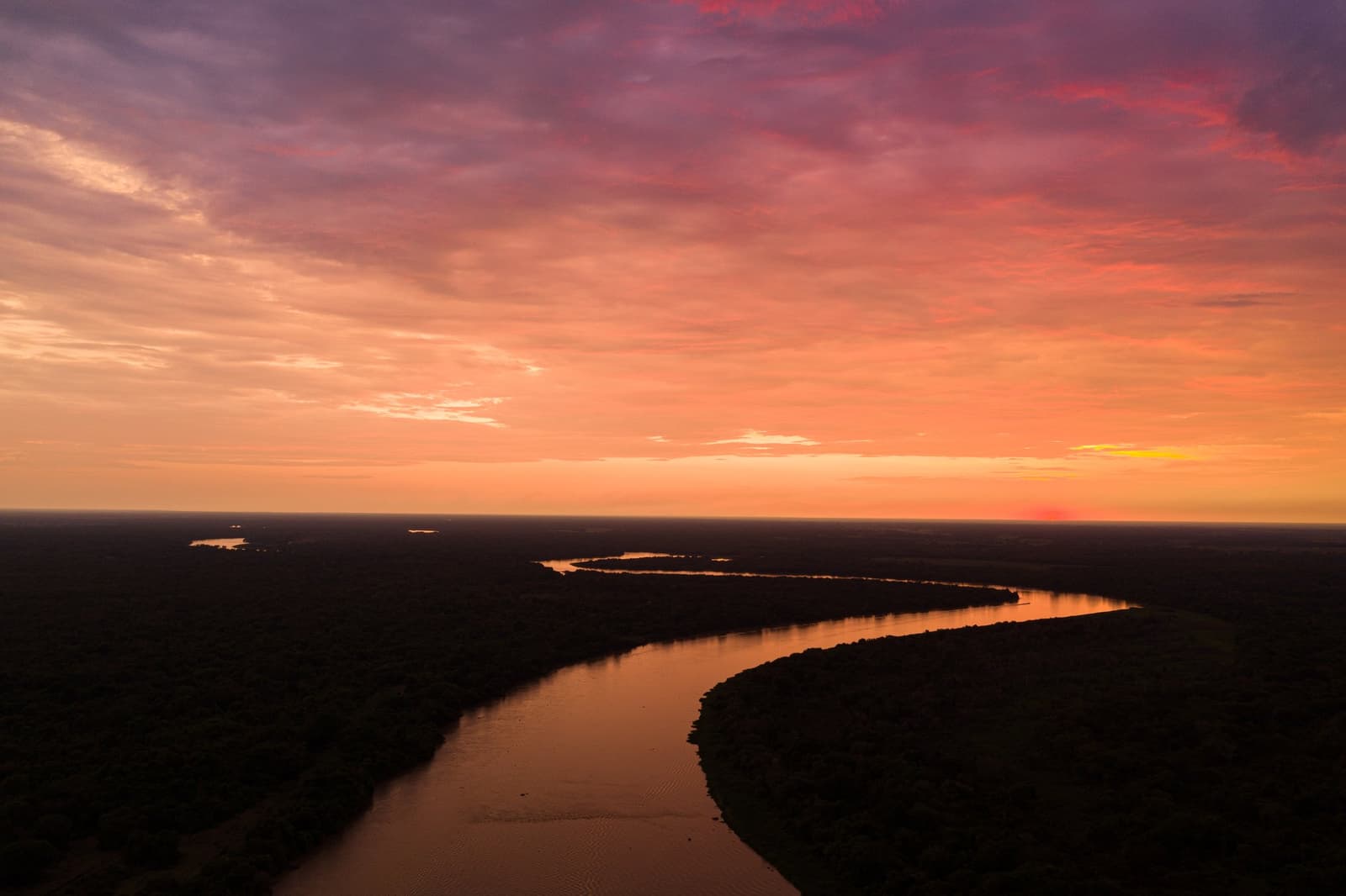 Laranja vivo, roxo profundo, a planície alagada sem fim. Photo: Roi Shomer Vista aérea de um rio sinuoso refletindo um céu de pôr do sol em tons de laranja e violeta intensos sobre extensas planícies alagadas no safari de onça-pintada no Pantanal Porto Jofre
