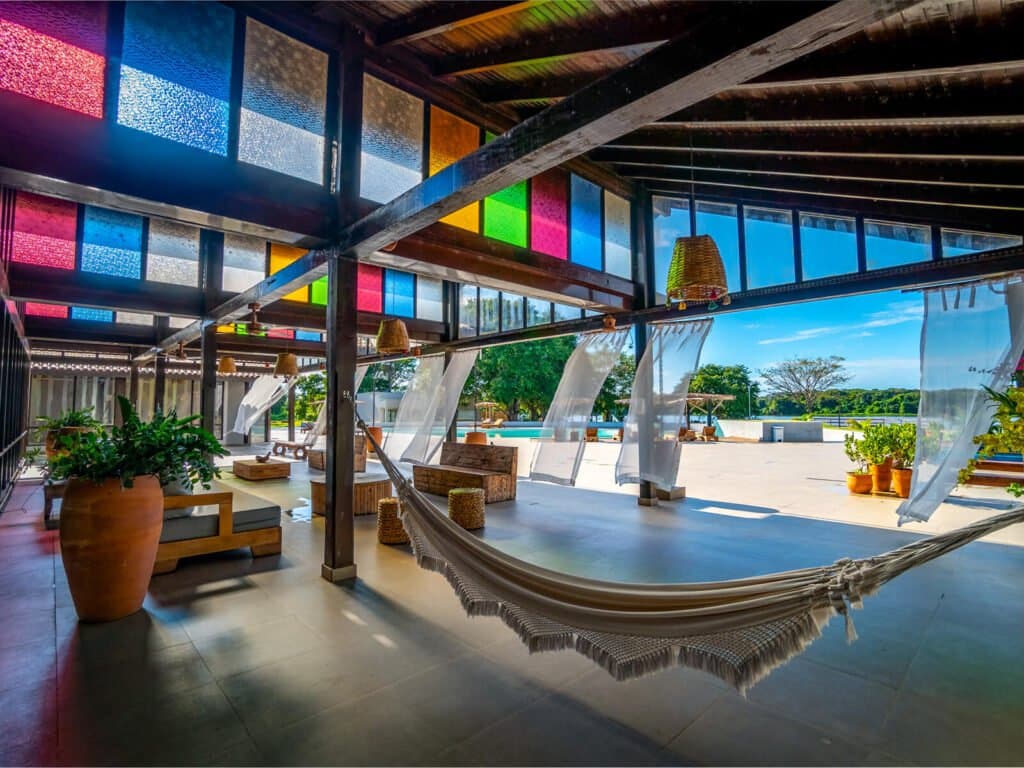 Rede balançando sob um teto de vidro colorido, o descanso do meio-dia na pousada tem charme próprio. Lounge aberto de pousada com rede de franja em primeiro plano, painéis de vidro coloridos no teto, luminárias de rattan e vista para o terraço ensolarado com piscina e vegetação ao fundo