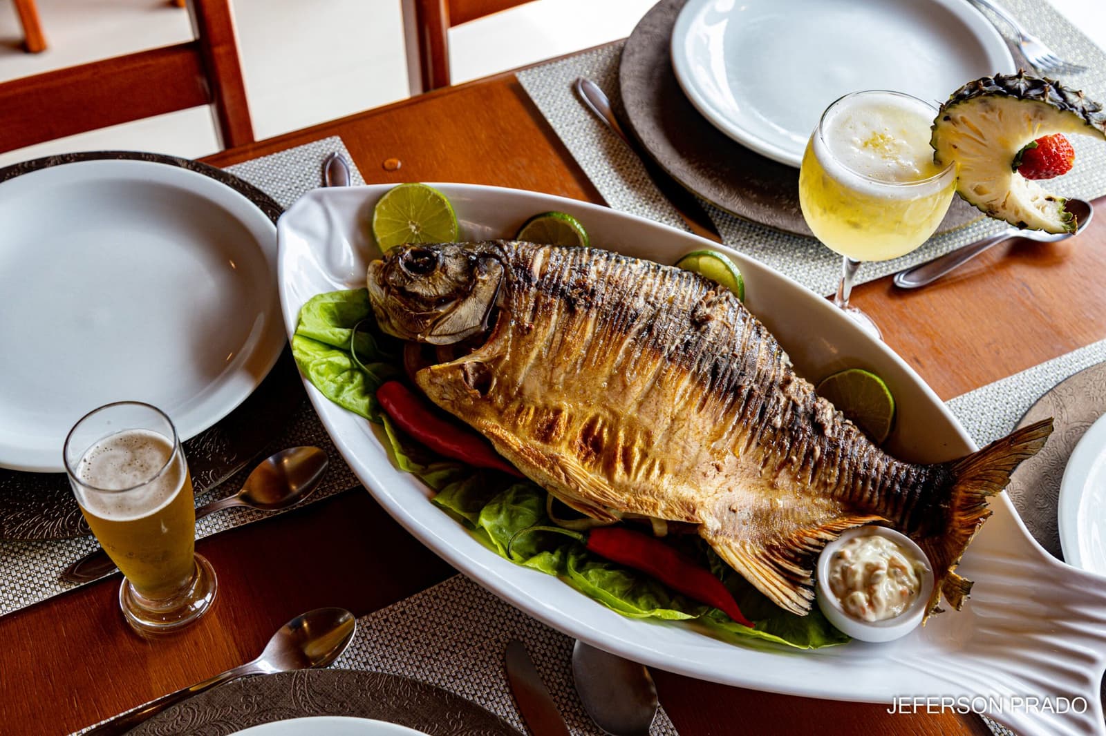 Peixe grelhado na hora, cerveja gelada, o almoço que a viagem merece. Photo: Jeferson Prado Um peixe inteiro grelhado servido em travessa branca com alface, pimentões vermelhos, rodelas de limão e molho, acompanhado de cerveja gelada e drinque de abacaxi numa mesa de jantar em pousada no Pantanal
