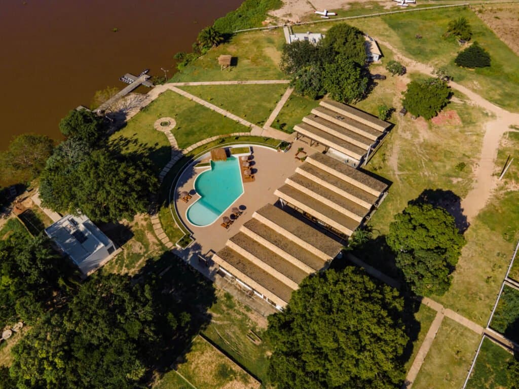 Piscina, deck no rio e mata em volta, a lodge vista de cima. Vista aérea de uma lodge de safari de onça-pintada no Pantanal em Porto Jofre, com piscina turquesa em formato orgânico, bangalôs com telhado de sapê, gramados cuidados e um deck privativo às margens do rio