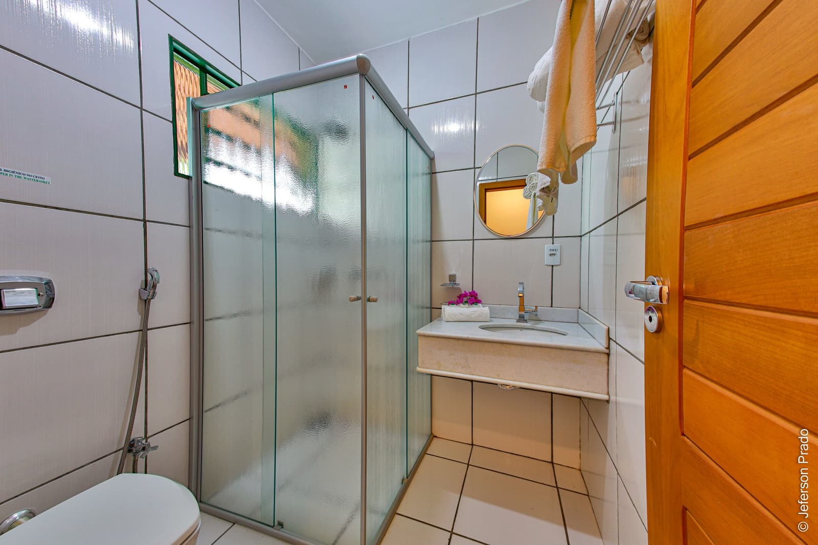 Toalhas frescas e chuveiro confortável para renovar as energias entre os safáris. Photo: Jeferson Prado Banheiro privativo limpo em uma pousada de Porto Jofre, com box de vidro, azulejos brancos, pia de mármore, espelho redondo e toalhas frescas no suporte aquecido