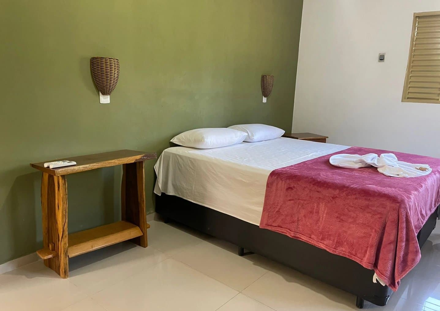 Conforto simples e aconchegante para descansar entre os safáris no rio. Quarto de lodge com cama de casal arrumada, cobertor vermelho e toalhas dobradas, paredes em tom verde-oliva e móveis rústicos de madeira