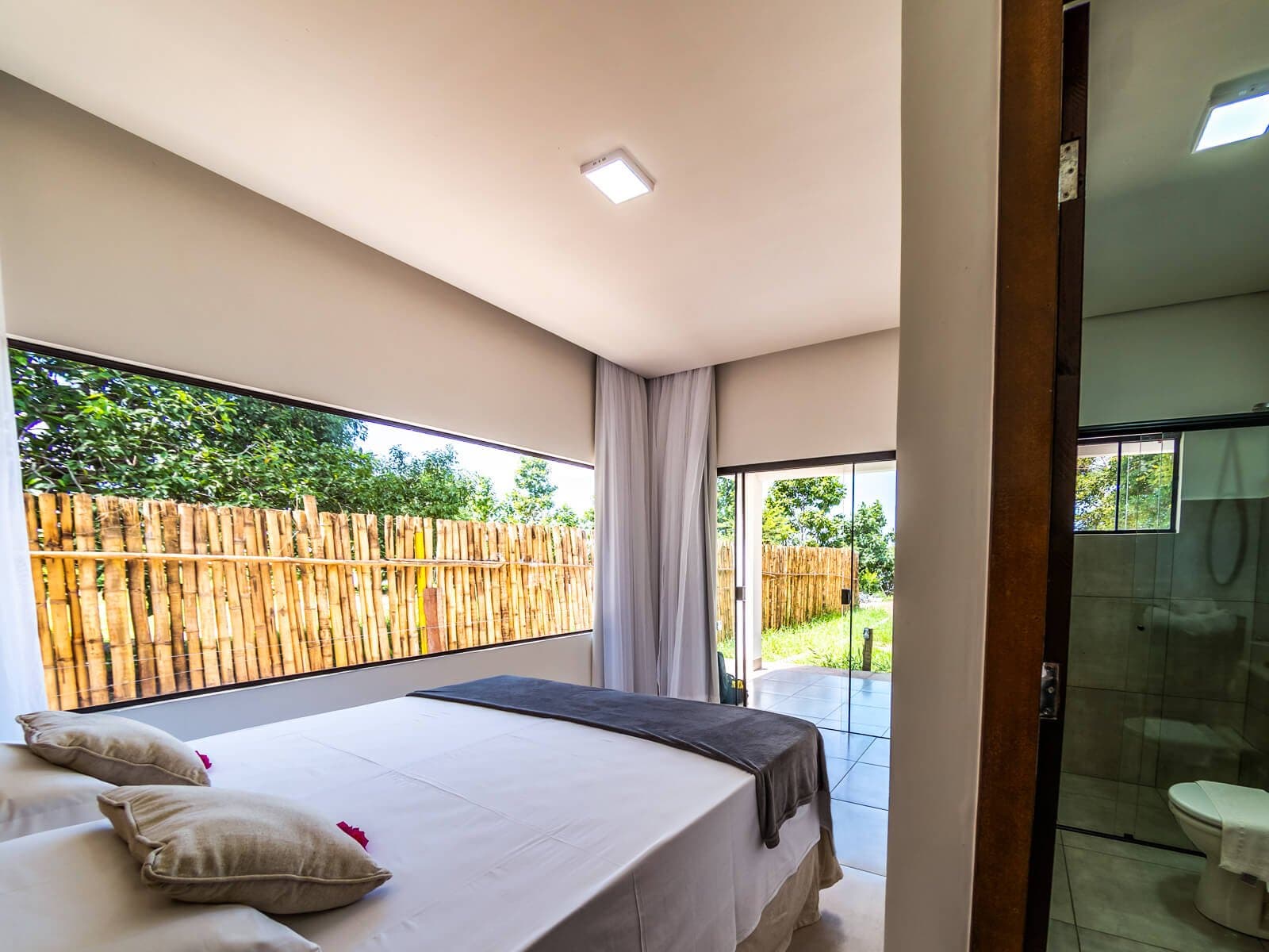 Conforto simples e natural, descanse bem antes de mais um amanhecer no rio. Quarto de casal iluminado em uma pousada de safari de onça no Pantanal em Porto Jofre, com roupa de cama branca, almofadas de linho, janela panorâmica com cerca de bambu e banheiro com parede de vidro