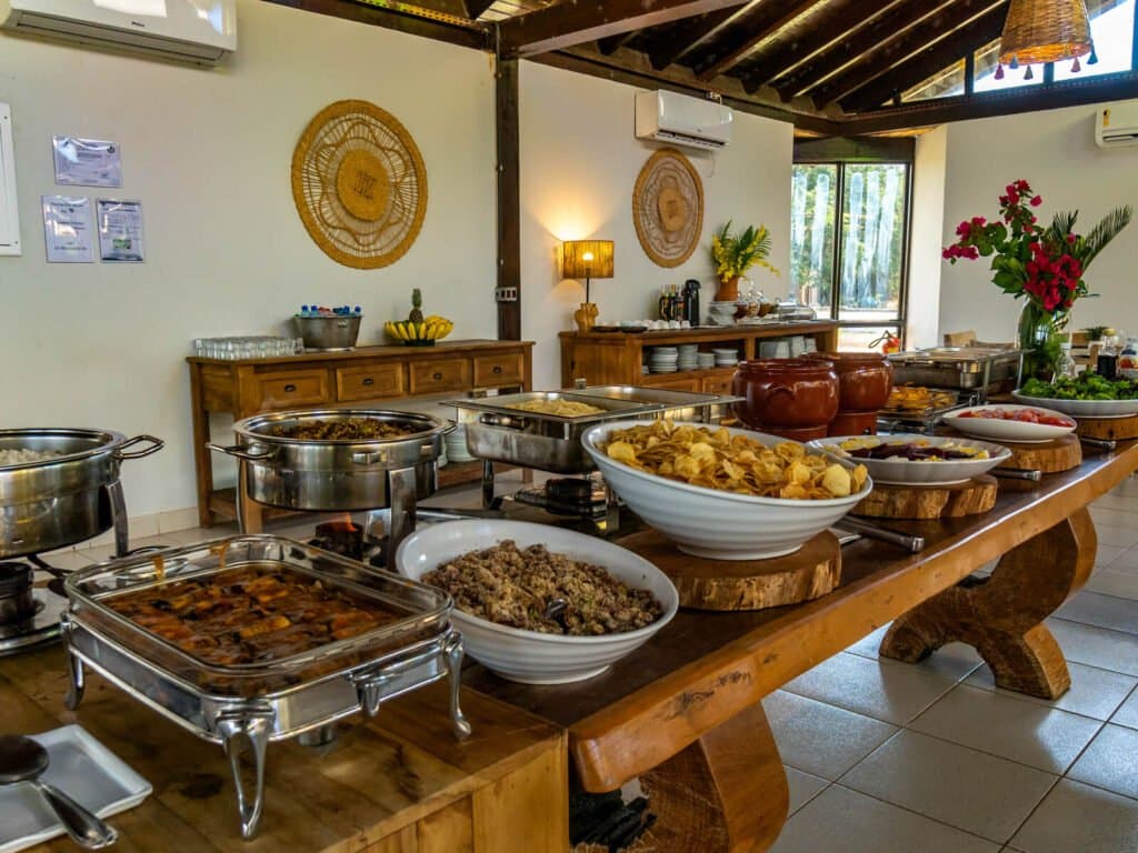 Culinária regional abundante para recarregar as energias entre os safáris. Um farto buffet brasileiro sobre uma mesa rústica de madeira em uma pousada de safari de onça-pintada no Pantanal de Porto Jofre, com rechauds, farofa, petiscos fritos e flores frescas