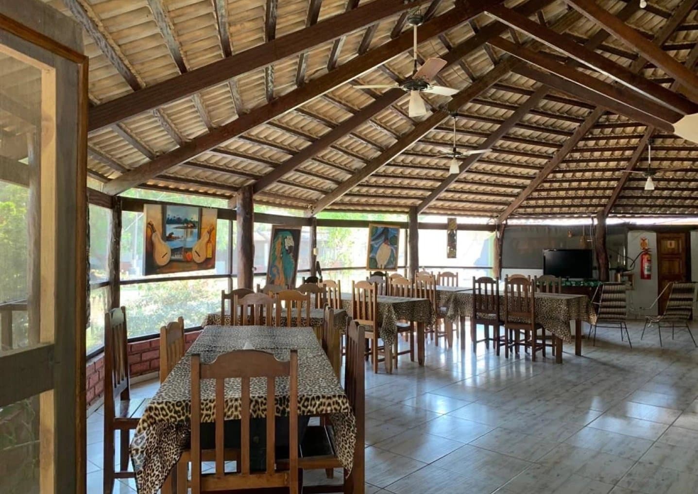 Vigas rústicas, arte da fauna e mesas prontas, o almoço também é parte do safari. Salão de refeições de lodge ao ar livre com teto alto de telha, vigas de madeira aparente, cadeiras de madeira, toalhas de mesa estampadas de onça e pinturas de fauna nas paredes teladas