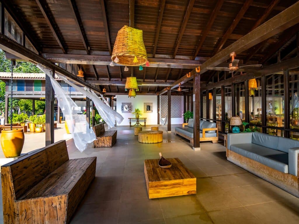 Madeira bruta, luminárias de palha e uma brisa que move as cortinas, o espaço perfeito para relaxar entre safaris. Lounge aberto de um lodge para safari de onça-pintada no Pantanal em Porto Jofre, com bancos rústicos de madeira maciça, luminárias de palha trançada e cortinas brancas ao vento