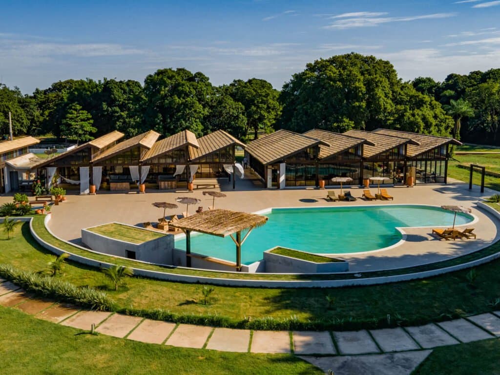 Palha, água fresca e verde por toda parte, a base perfeita para um safari inesquecível. Vista aérea de uma lodge de safari de onça no Pantanal com piscina curva de água turquesa, pavilhões com telhado de palha, espreguiçadeiras e gramados verdes entre árvores nativas