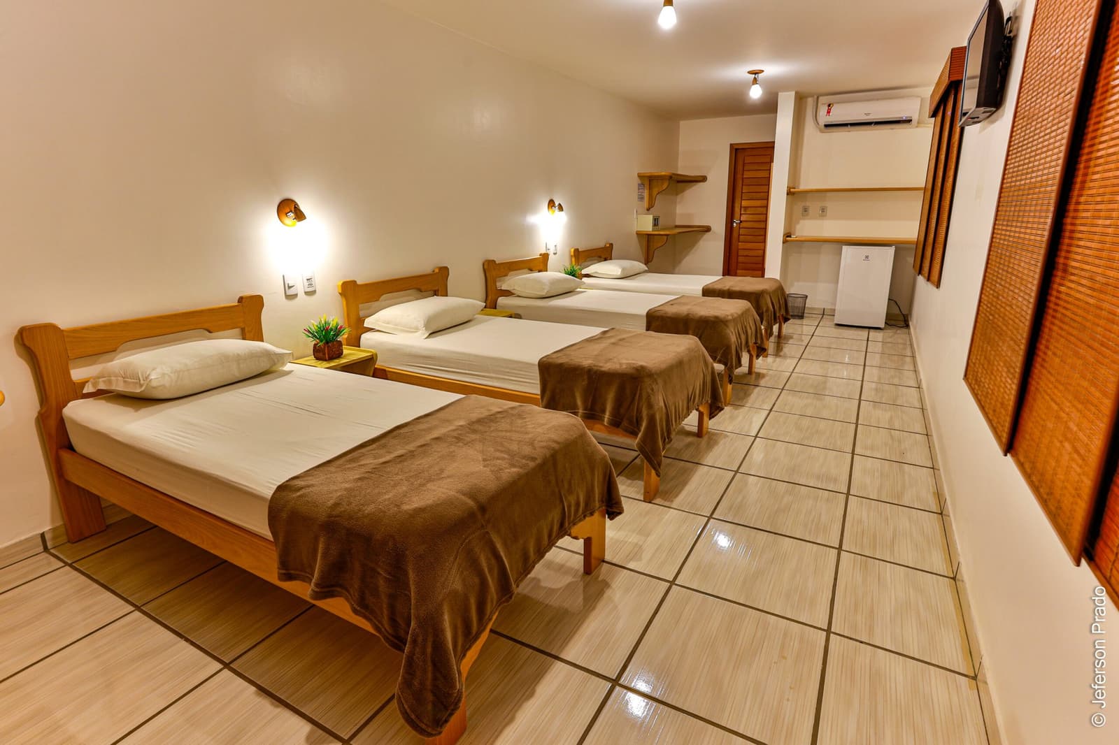 Conforto simples e aconchegante para descansar entre os safaris. Photo: Jeferson Prado Quarto quádruplo espaçoso em pousada de Porto Jofre para um safari de onças no Pantanal, com quatro camas de solteiro em madeira, roupas de cama brancas e mantas marrons, arandelas, ar-condicionado e frigobar