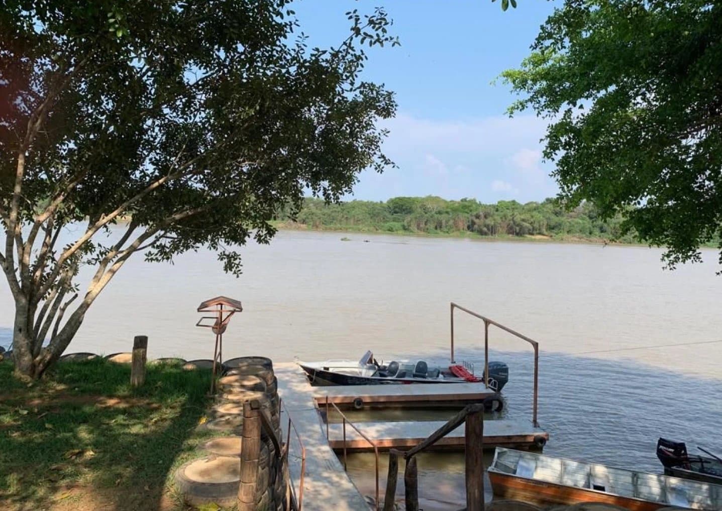 Barcos prontos, rio à espera, o embarque para o Pantanal começa aqui. Um deck de madeira com barcos a motor atracados em um amplo rio de águas barrentas, emoldurado por árvores ribeirinhas em um dia ensolarado