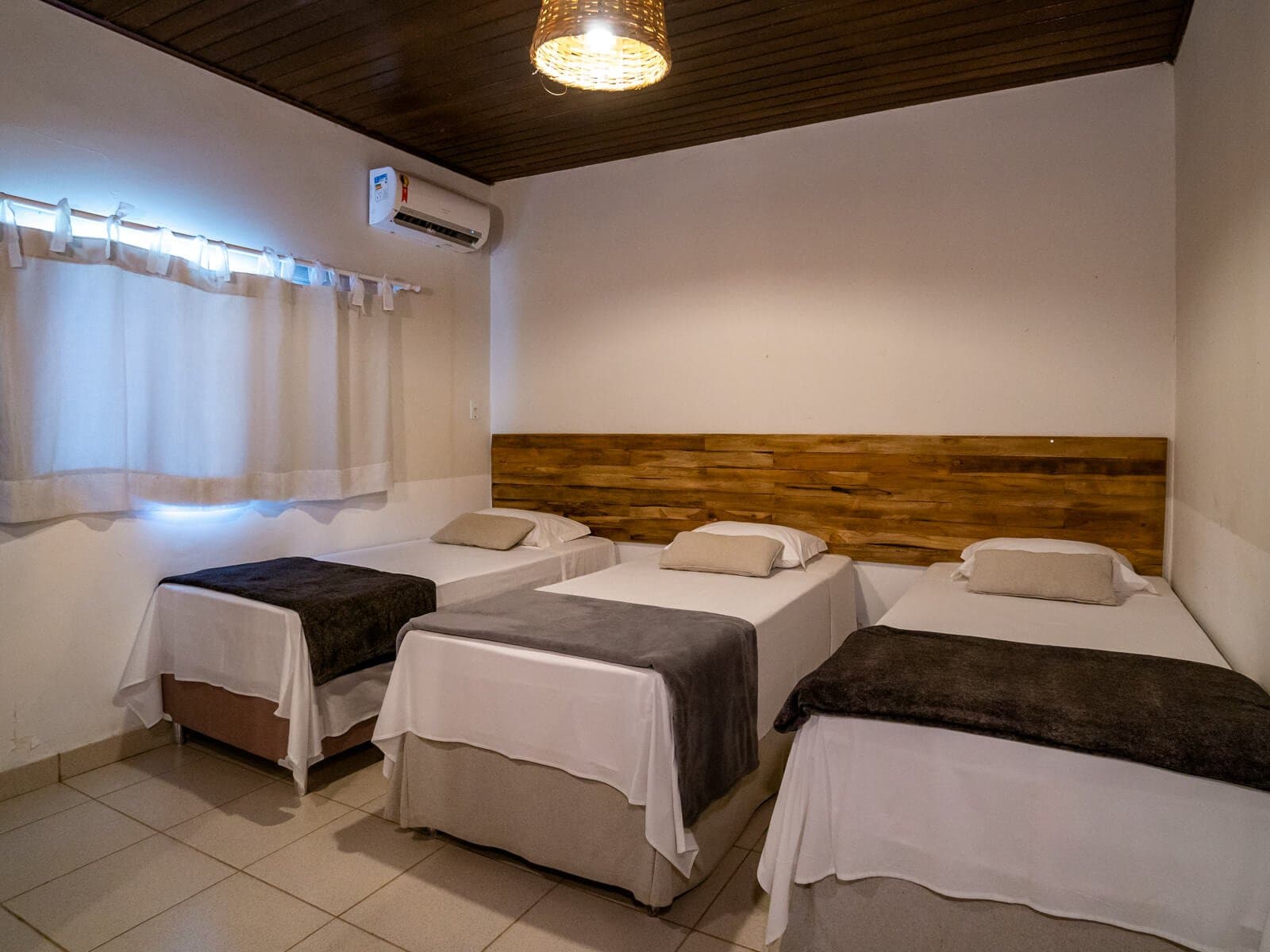 Conforto simples e aconchegante para recarregar as energias entre os safáris. Quarto triplo arrumado em uma pousada em Porto Jofre com três camas de solteiro com roupas de cama brancas, cabeceira de madeira rústica, ar-condicionado e lustre de vime