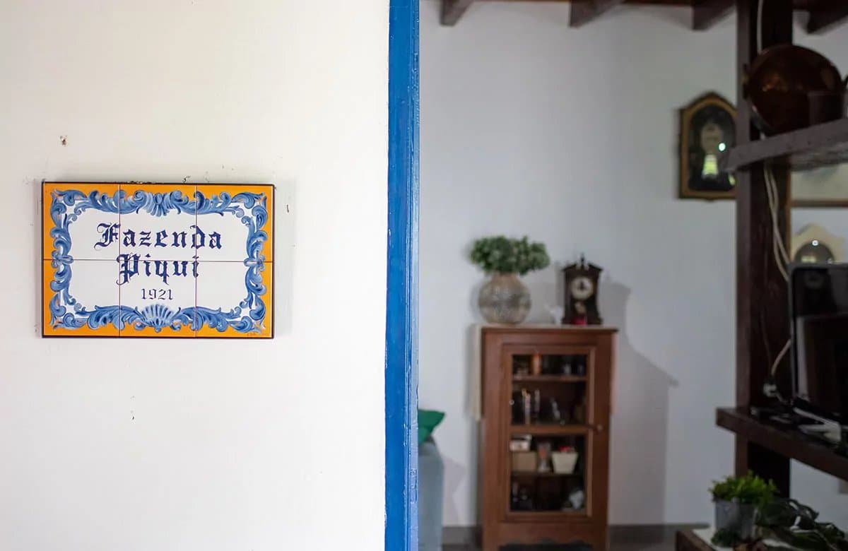 Desde 1921, o azulejo conta a história sem precisar de palavras. Placa de azulejo pintado à mão com os dizeres 'Fazenda Piqui 1921' fixada em parede branca ao lado de uma moldura de porta azul que dá acesso a um interior rústico