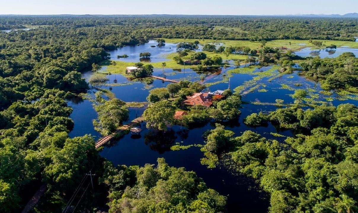 Na cheia, a pousada vira ilha. Quem passa por isso não esquece a vista. Vista aérea de um ecolodge remoto cercado por áreas úmidas alagadas e densa floresta verde em um passeio pelo Pantanal a partir de Campo Grande, com vitórias-régias pontilhando os canais escuros e telhados vermelhos ligados por passarelas de madeira
