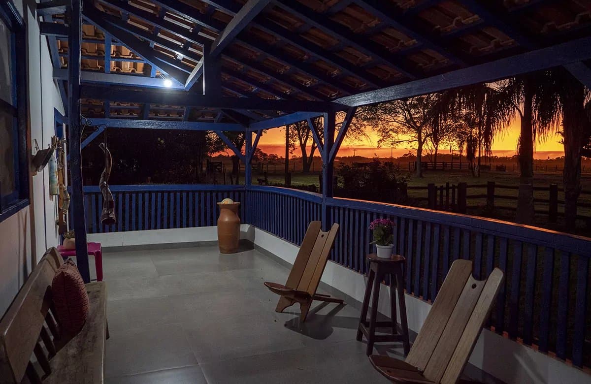 Fim de tarde na pousada: simples assim, difícil de esquecer. Varanda coberta com gradil de madeira pintado de azul e cadeiras rústicas com vista para uma paisagem aberta iluminada por um pôr do sol laranja intenso em um tour pelo Pantanal saindo de Campo Grande