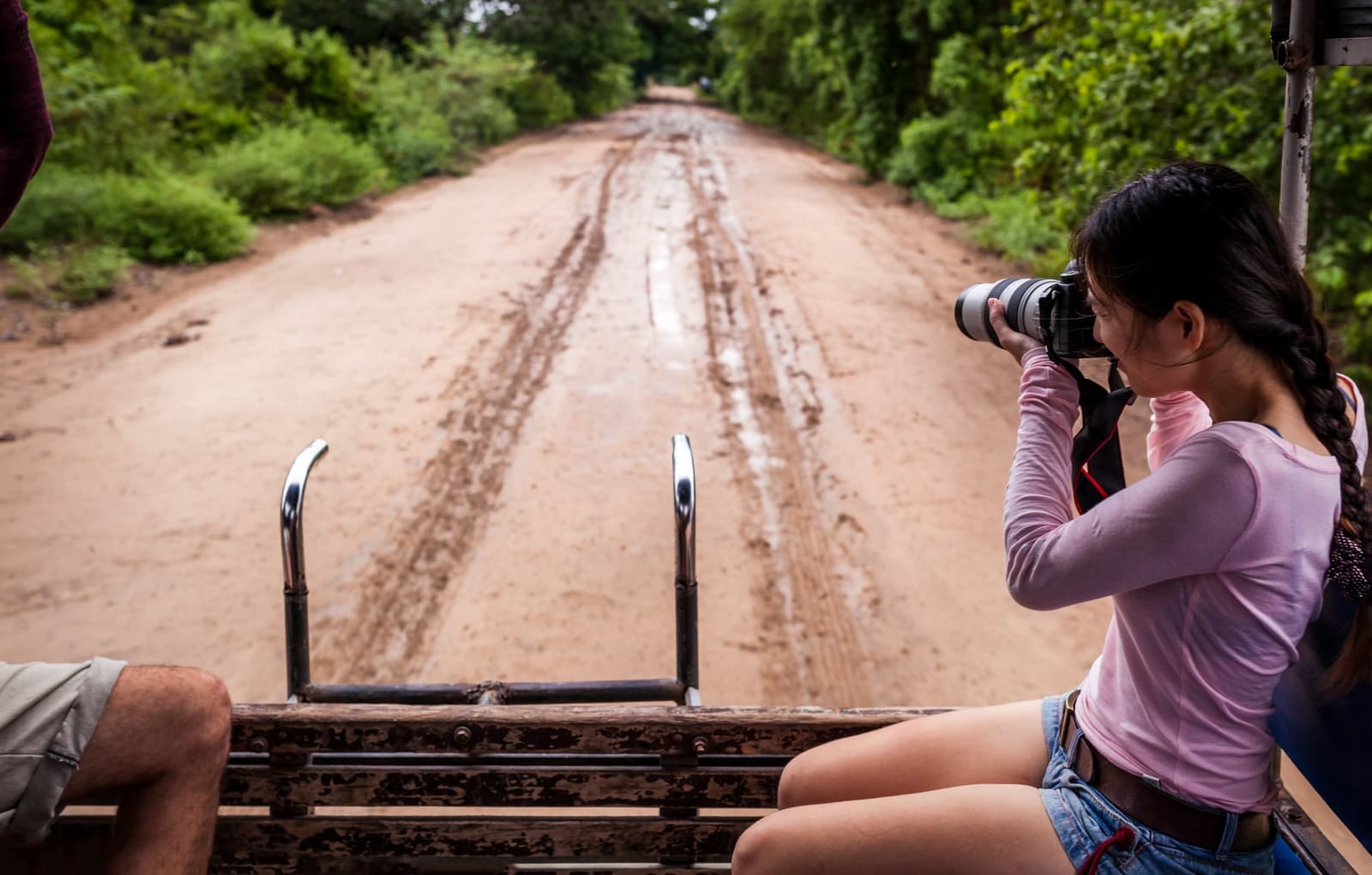 Lama na estrada é sinal de que a aventura começou de verdade. Uma viajante fotografa com uma lente teleobjetiva a partir da caçamba aberta de um 4x4 em uma estrada de terra vermelha ladeada por vegetação verde durante um passeio pelo Pantanal saindo de Campo Grande