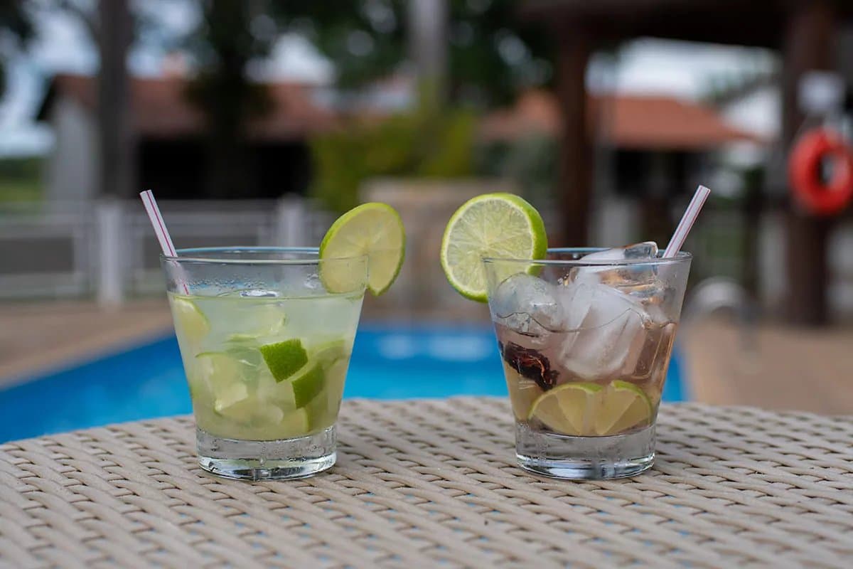 Uma caipirinha merece outra, só depois de chegar do passeio. Dois drinques gelados com limão e canudos sobre mesa de vime ao lado de uma piscina azul em uma ecolodge do Pantanal