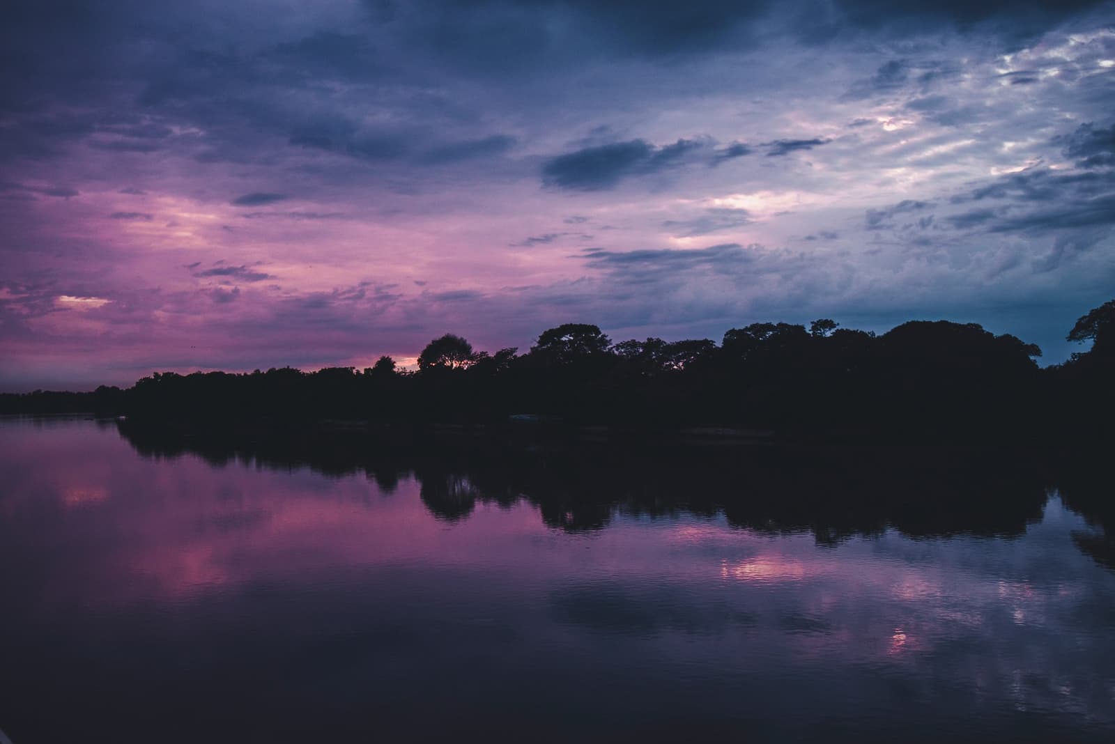 O Pantanal não economiza no fim do dia, o céu e o rio dividem o palco. Céu de pôr do sol em tons de roxo e rosa refletido em um rio calmo, com silhueta escura de árvores no horizonte