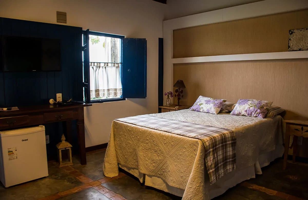 Conforto sem frescura, do jeito que o Pantanal pede. Quarto de casal aconchegante em pousada para o passeio no Pantanal saindo de Campo Grande, com colcha bordada, almofadas floridas em lilás, janelas de madeira azul abertas para a vegetação e escrivaninha de madeira