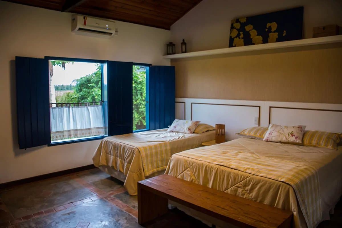 A simplicidade que cabe bem aqui: cama boa, janela aberta, natureza lá fora. Quarto de pousada com duas camas de roupa xadrez amarela, piso de cerâmica, teto de madeira e janelas com venezianas azuis abertas para vegetação verde