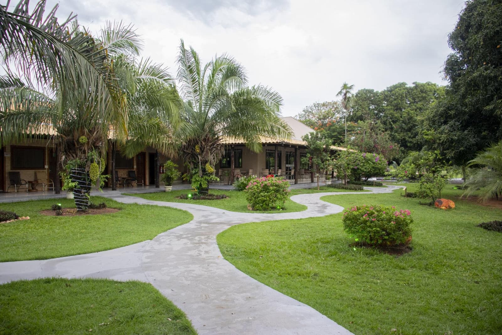 The kind of garden that makes you slow your pace before you even try. Winding concrete pathways curve through a lush tropical garden leading to low-slung thatched-roof lodge buildings on a Pantanal Tour from Cuiabá, with palm trees, flowering shrubs, and manicured lawns under a cloudy sky