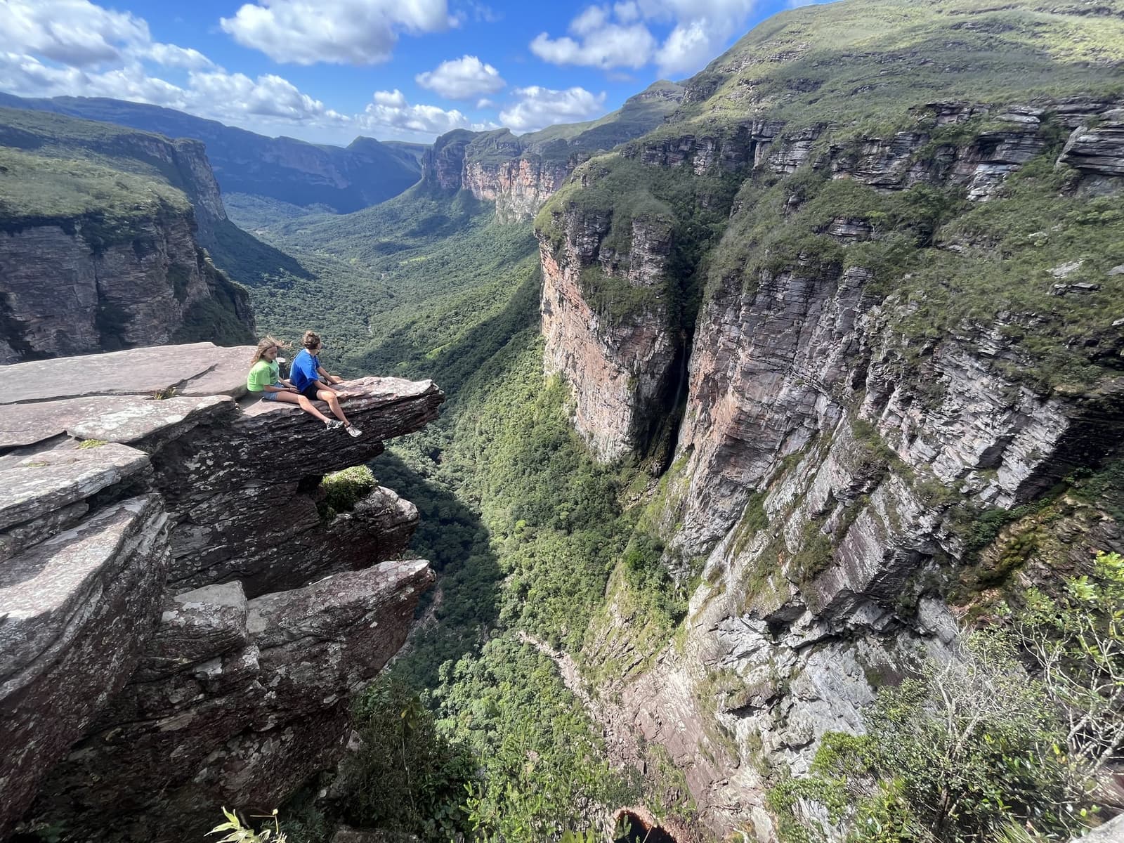 Ce belvédère se mérite. La vallée du Pati sait garder ses secrets. Deux randonneurs assis au bord d'un promontoire rocheux surplombent un canyon profond aux falaises de grès verticales et à la végétation luxuriante dans la Chapada Diamantina