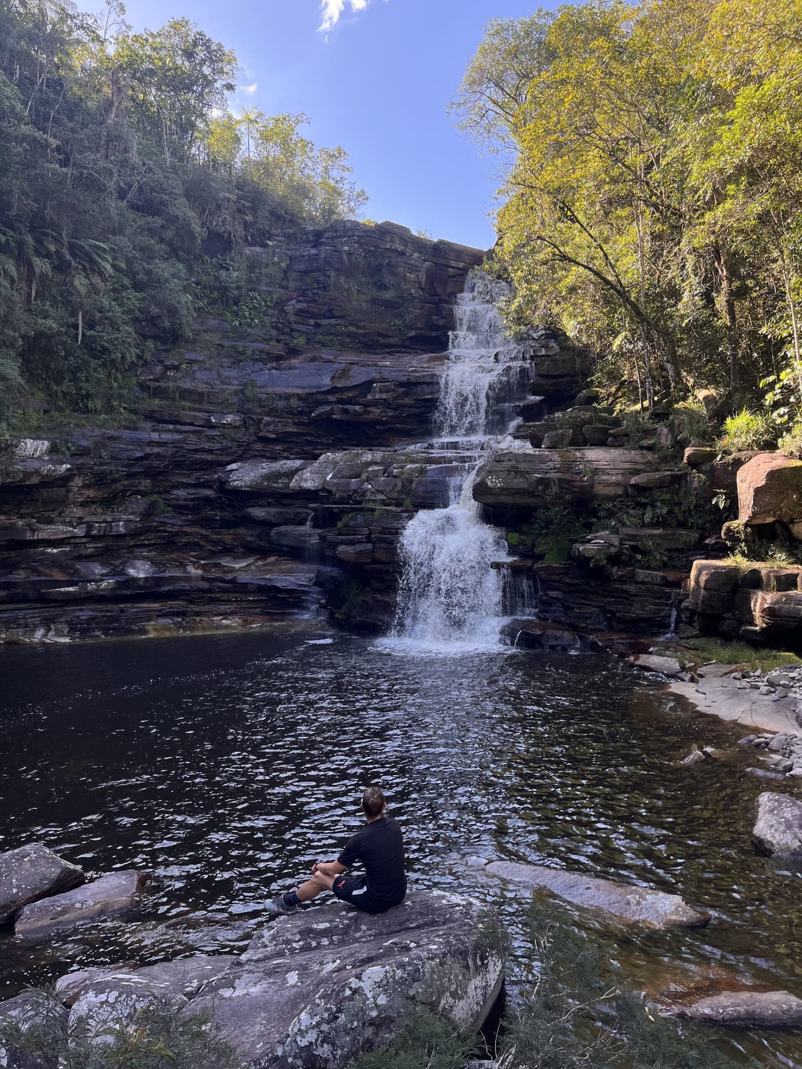 Le genre d'endroit où on oublie complètement l'heure. Un randonneur assis seul sur un rocher plat de grès au bord d'un bassin naturel sombre contemple une cascade en gradins dégringolant sur des parois rocheuses en couches lors de la randonnée dans la Pati Valley, entouré d'une forêt verdoyante sous un ciel bleu