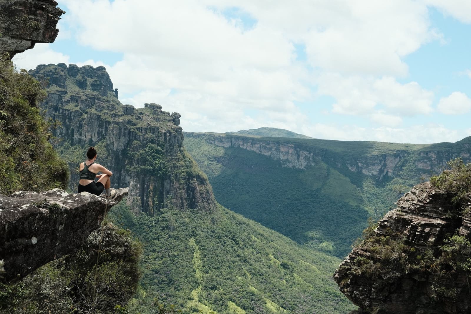 Ce genre de silence, il faut le mériter à pied. Une randonneuse assise seule sur une corniche rocheuse contemple un vaste canyon verdoyant lors de la randonnée dans la vallée de Pati, avec de hautes falaises de grès et des versants boisés s'étendant à perte de vue sous un ciel partiellement nuageux
