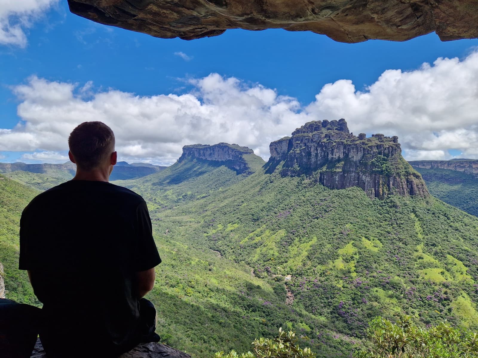 Ce cadre, c'est la roche qui l'a sculpté. La vue, elle se mérite. Un randonneur assis sous un surplomb rocheux contemple le mesa imposant du Morro do Castelo se dressant au-dessus de collines verdoyantes lors de la randonnée dans la Pati Valley