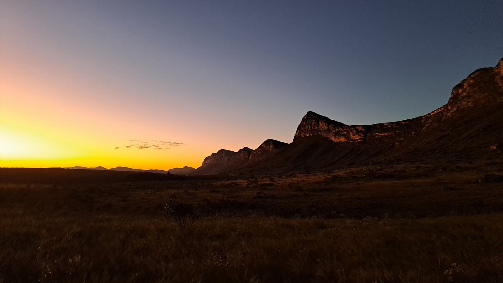 Le soleil passe, la roche reste. La Chapada sait très bien se faire remarquer. Un coucher de soleil aux teintes orange et jaune illumine l'horizon derrière la silhouette d'un escarpement rocheux lors de la randonnée dans la vallée de Pati, avec une savane ouverte au premier plan