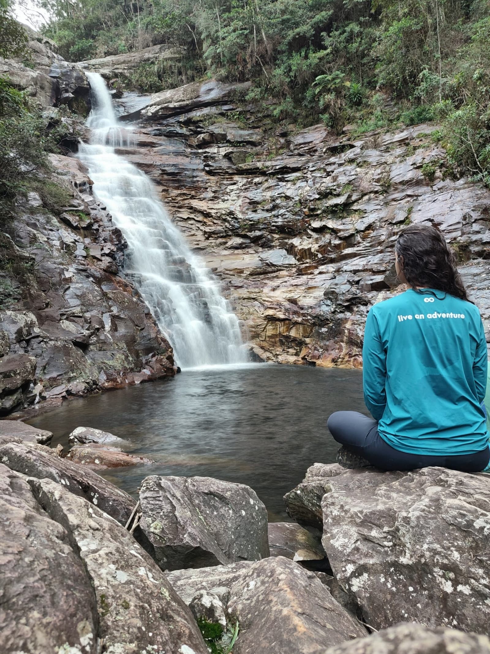 Certaines pauses valent plus que mille pas. Une randonneuse en haut sarcelle est assise en tailleur sur des rochers et contemple une cascade soyeuse se déversant dans un bassin tranquille encadré de falaises stratifiées et de forêt
