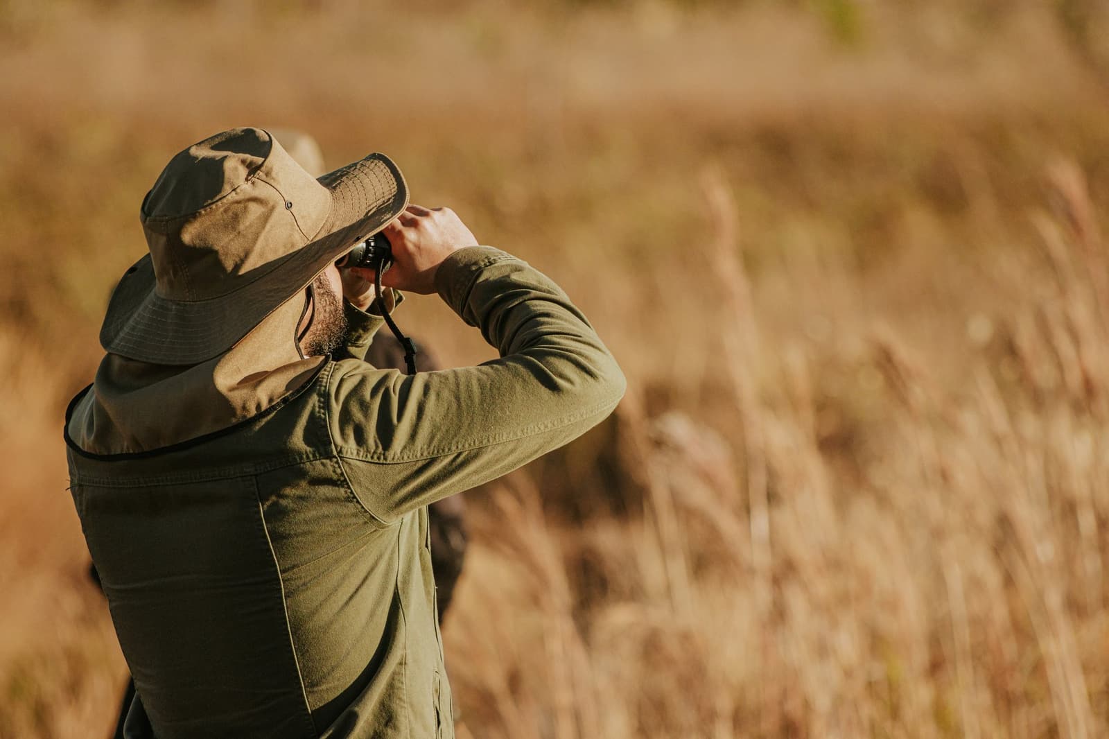 Campo alto, binóculo firme, e a lista de aves crescendo sem pressa. Um viajante barbudo com chapéu de abas largas usa binóculos para observar o campo cerrado dourado durante o birdwatching na Pousada Trijunção