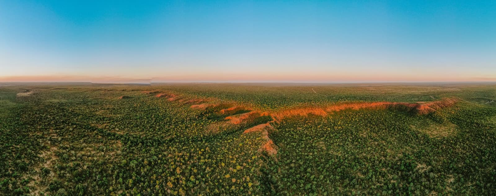O Cerrado vai até o fim do mundo, e aquela faixa vermelha no meio lembra que a terra tem estilo próprio. Vista aérea panorâmica da paisagem de Cerrado da Pousada Trijunção ao pôr do sol, com uma crista de laterita vermelha cortando a vegetação verde sob um céu em tons de rosa e azul pastel