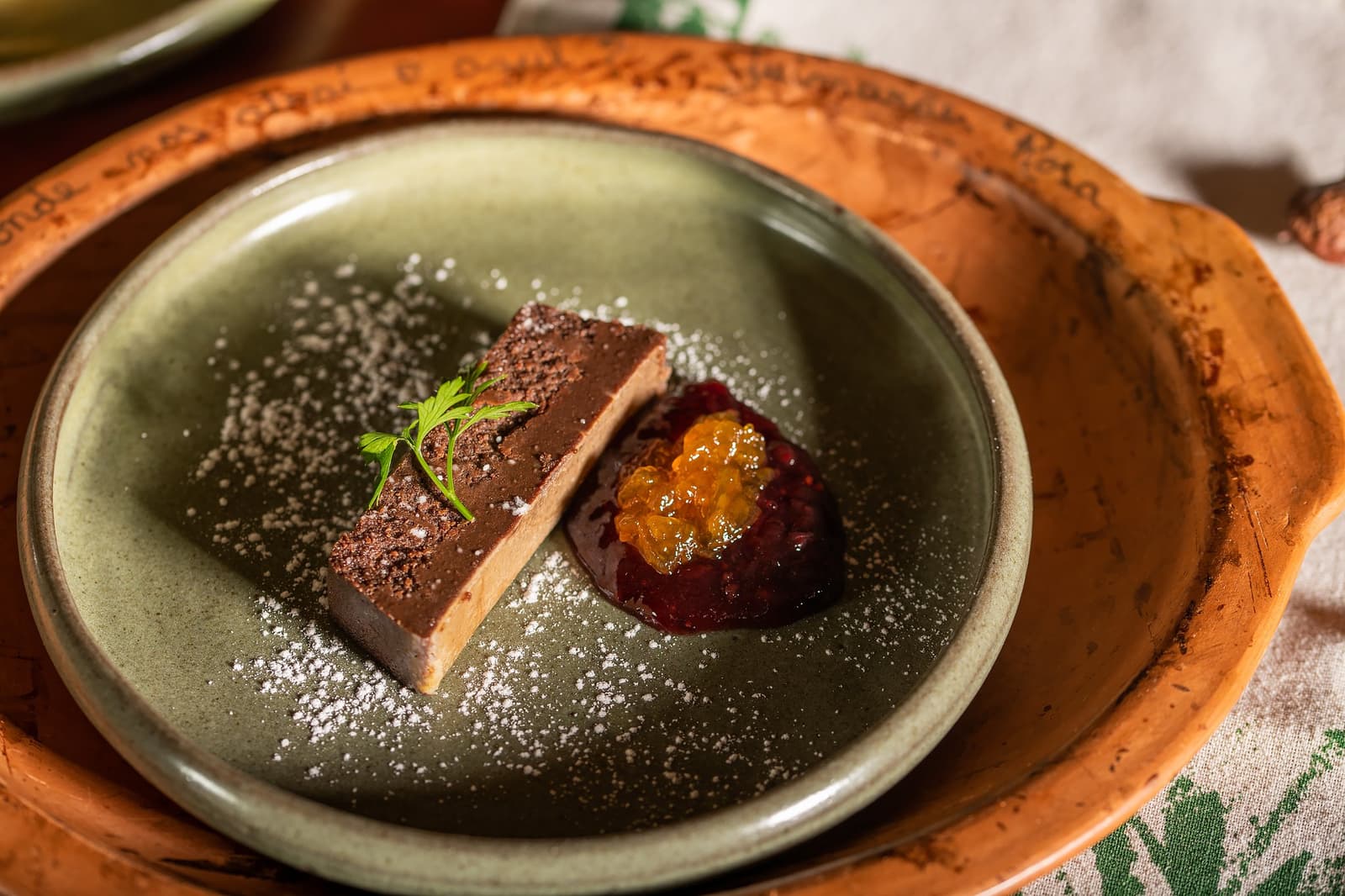 No Cerrado, até a sobremesa conta uma história do lugar. Fatia de terrine de chocolate polvilhada com cacau e açúcar de confeiteiro, acompanhada de compota de frutas com pérolas douradas em prato de cerâmica verde-sálvia na Pousada Trijunção