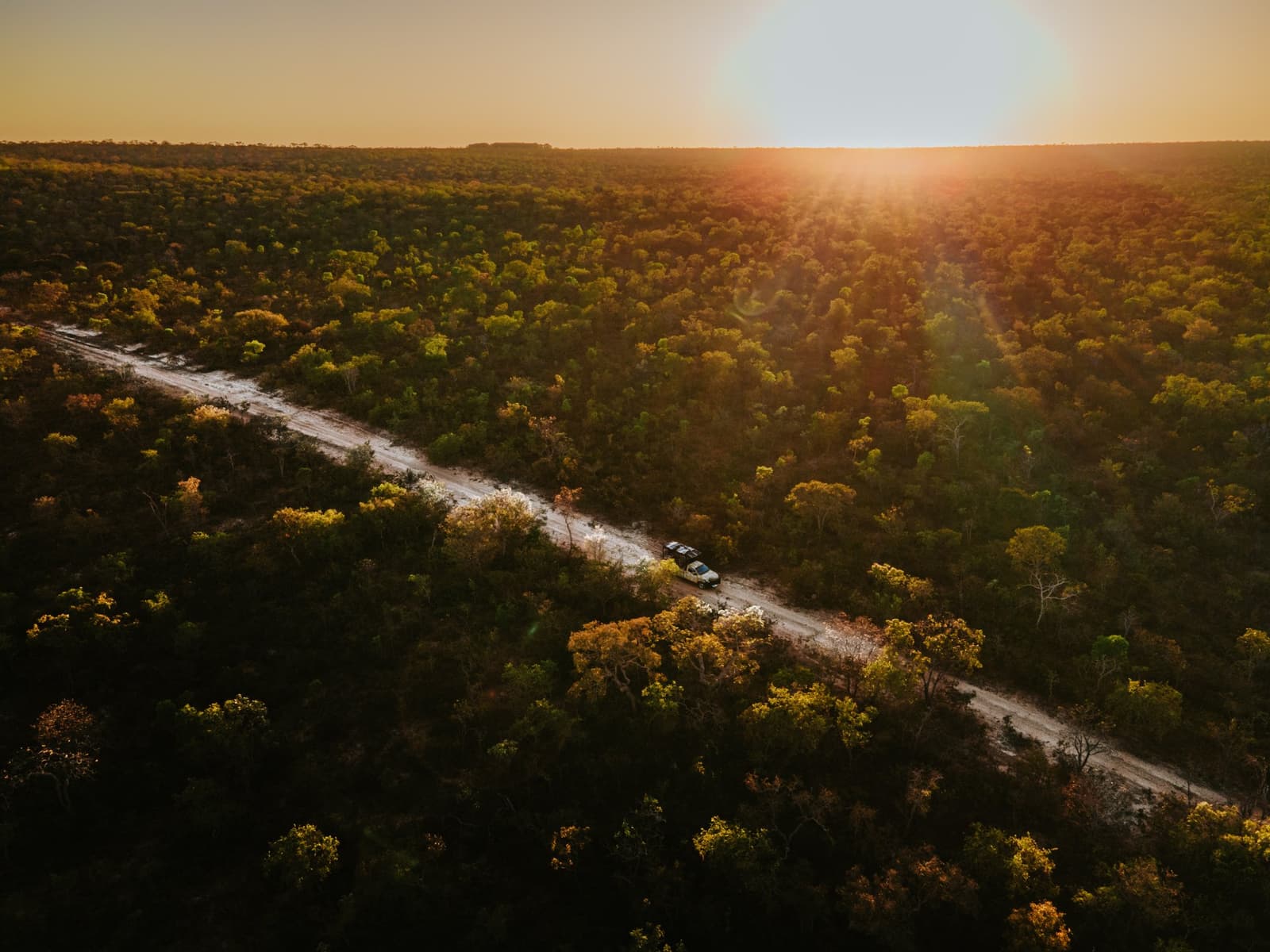 Estrada de chão, mata fechada e o sol despedindo-se do dia com tudo. Vista aérea de dois veículos 4x4 em uma estrada de terra cortando o cerrado denso da Pousada Trijunção, banhados pela luz dourada do pôr do sol