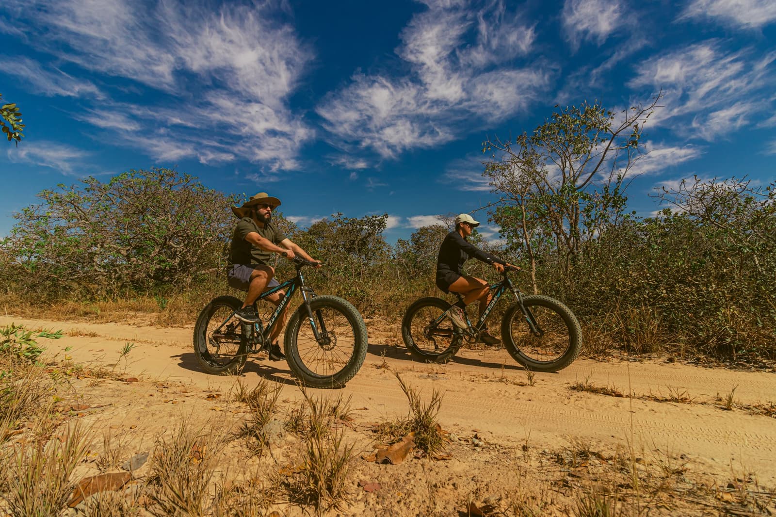 Pneus largos, areia fina e um Cerrado que não deixa ninguém parado. Dois ciclistas pedalando fat bikes em uma trilha de areia no Cerrado da Pousada Trijunção, cercados pela vegetação nativa sob um céu azul intenso