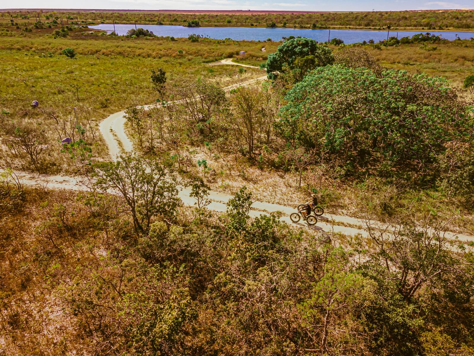 A trilha serpenteia pelo Cerrado e a lagoa aparece logo ali na frente. Vista aérea de dois ciclistas em fat bikes percorrendo uma trilha de terra sinuosa pelo cerrado da Pousada Trijunção, com uma lagoa azul visível ao fundo
