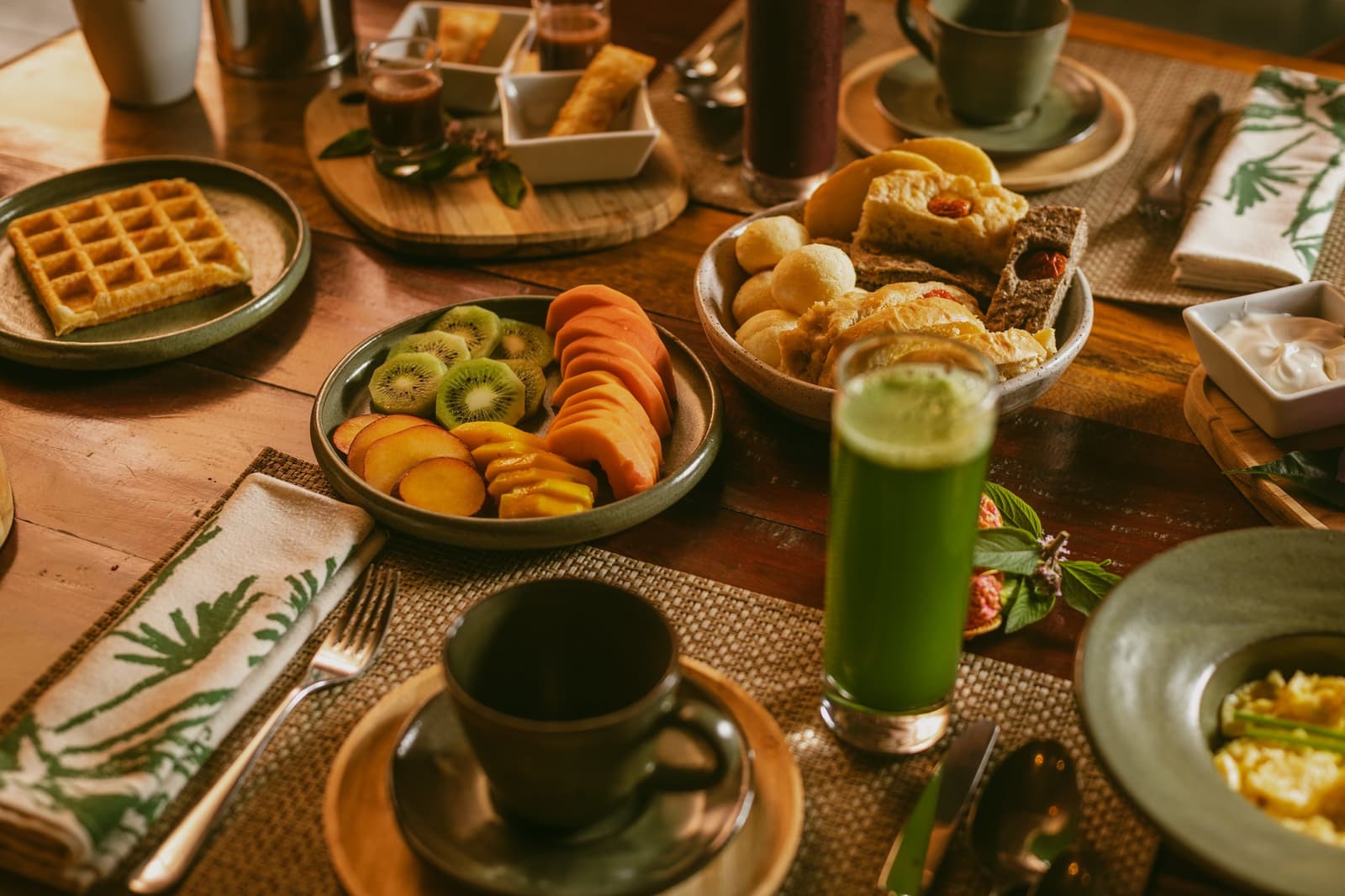 Pão de queijo, suco verde e frutas frescas. O Cerrado começa bem antes da trilha. Mesa de café da manhã farta na Pousada Trijunção, com fatias de kiwi, pêssego e mamão, waffles dourados, pãezinhos quentinhos, suco verde vibrante e uma caneca de café em cerâmica sobre jogo americano trançado