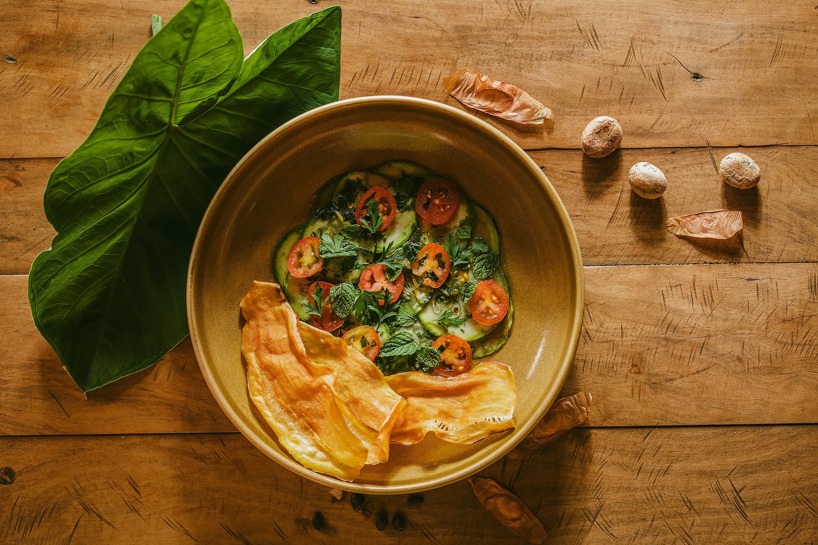 Cozinha de pousada que cheira a mata e sabe a casa. Vista aérea de um bowl de cerâmica com abobrinha fatiada, tomates-cereja e ervas frescas acompanhados de chips crocantes na Pousada Trijunção, cercado por folhas tropicais e sementes silvestres sobre uma mesa de madeira rústica