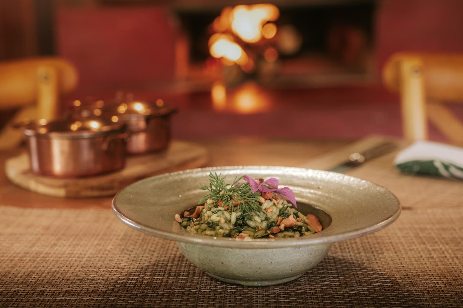 A lareira aquece, a comida cuida do resto. Uma tigela de cerâmica artesanal com risoto de ervas decorado com endro fresco e uma flor comestível rosa, servido à beira da lareira da Pousada Trijunção com panelas de cobre brilhando ao fundo