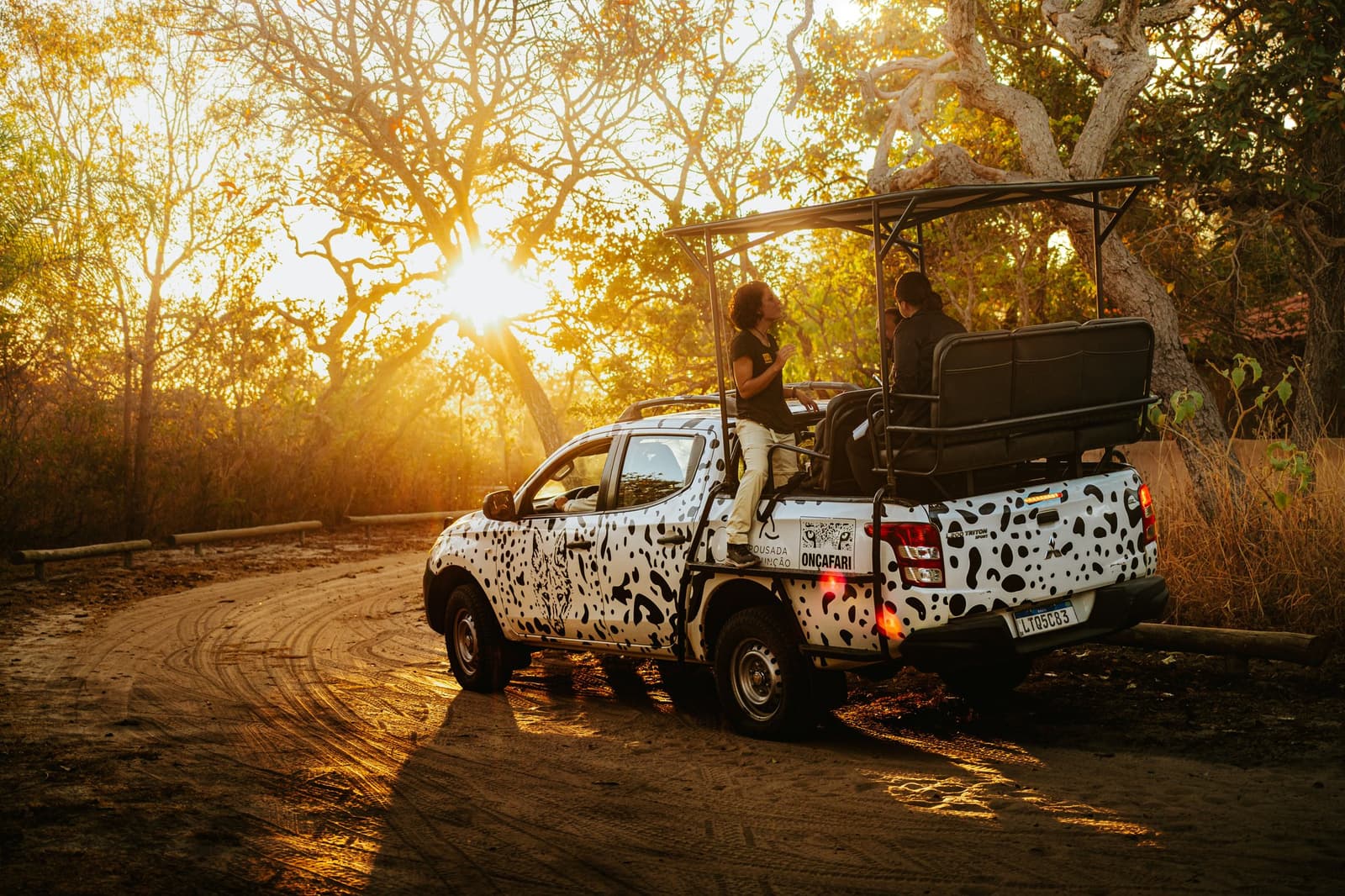 O melhor momento para trocar ideia? No fim do dia, com a poeira ainda no ar. Veículo safari da Onçafari com estampa de onça e hóspedes a bordo parado em estrada de terra durante pôr do sol dourado no Cerrado da Pousada Trijunção