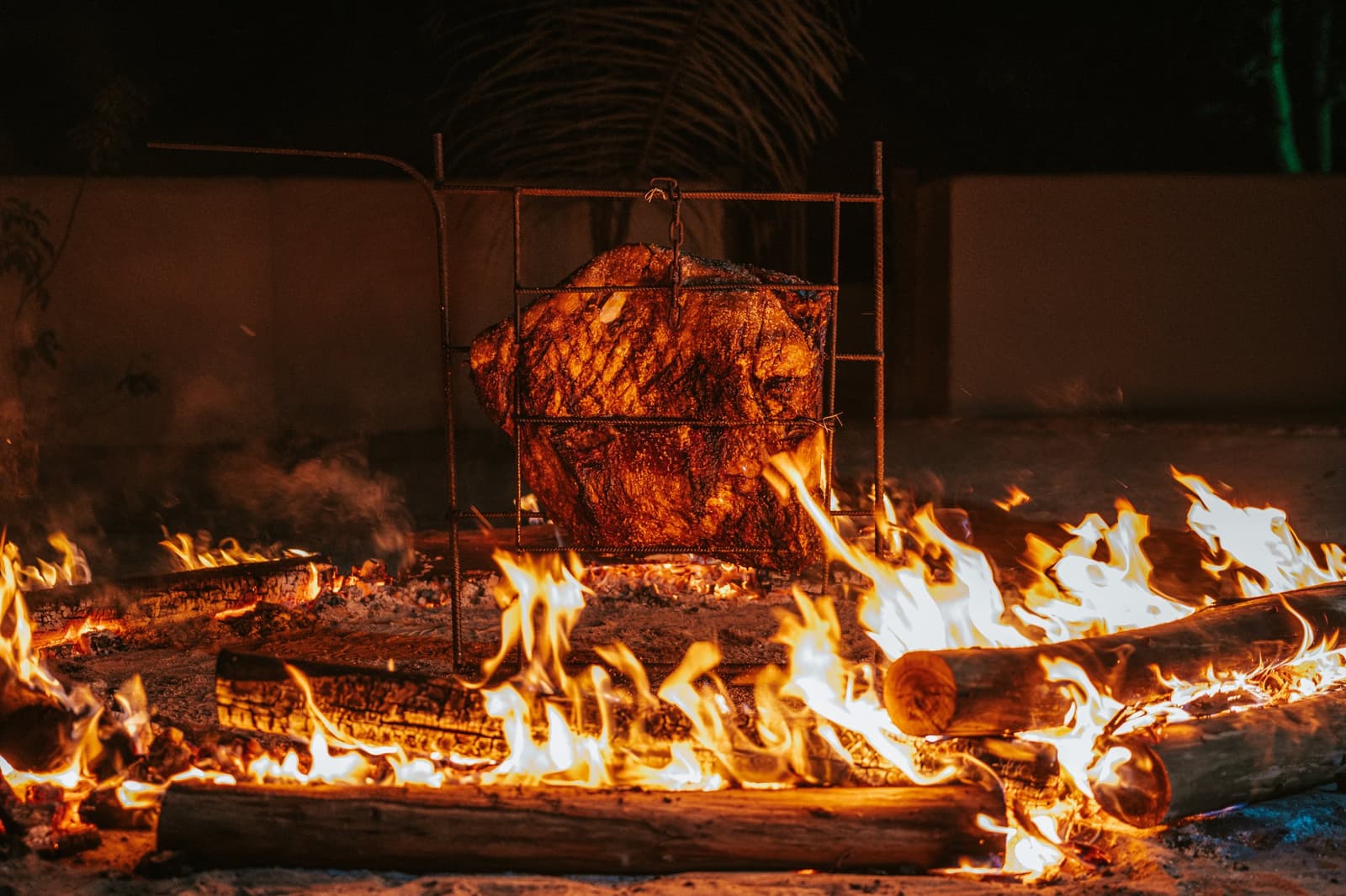Fogo de verdade, lenha de verdade, e tempo suficiente para fazer direito. Um grande corte de carne assando lentamente em uma grelha de ferro rústica sobre chamas de lenha à noite na Pousada Trijunção, no coração do Cerrado brasileiro