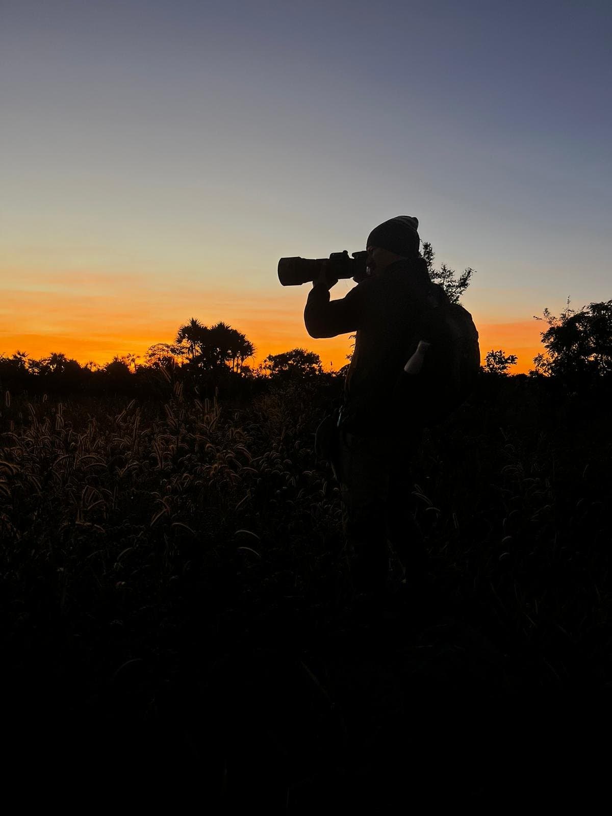 A luz dura poucos minutos. Ainda bem que alguém estava pronto. Silhueta de um fotógrafo de vida selvagem com uma lente telephoto apontada ao horizonte, entre as gramíneas do Cerrado sob um céu alaranjado do amanhecer na Pousada Trijunção